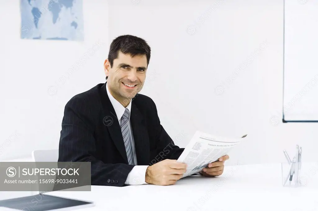 Businessman sitting at desk in office, reading newspaper, smiling at camera