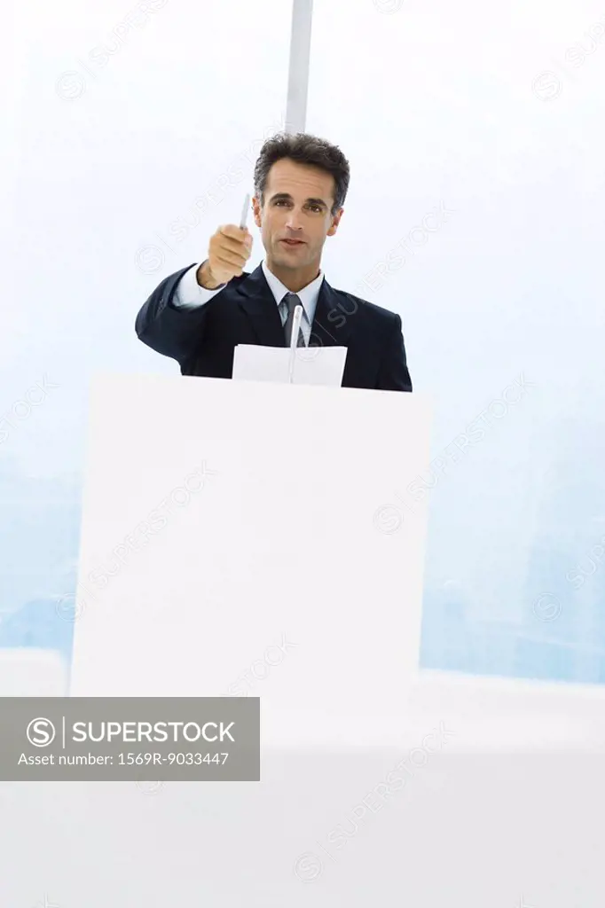 Executive standing behind lectern, pointing with pen at camera