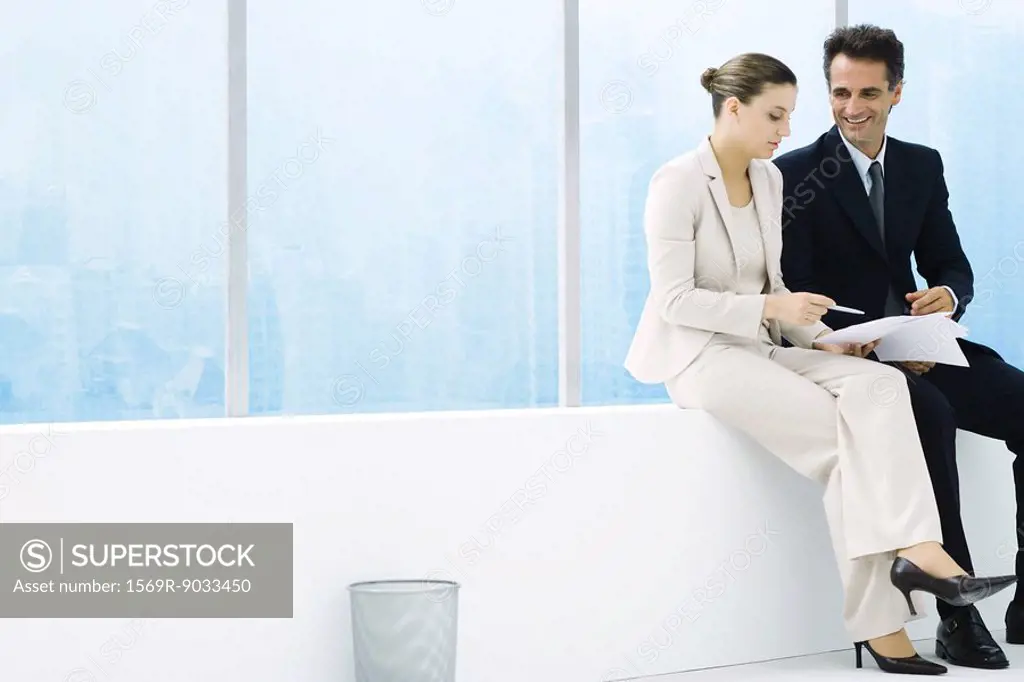 Male and female executives sitting on ledge by window, discussing document