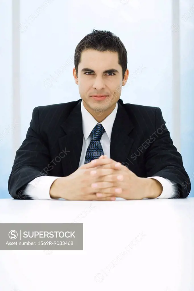 Businessman sitting at table with hands clasped, looking at camera