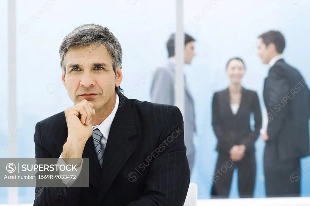 Graying businessman with hand under chin, looking away, colleagues chatting in background