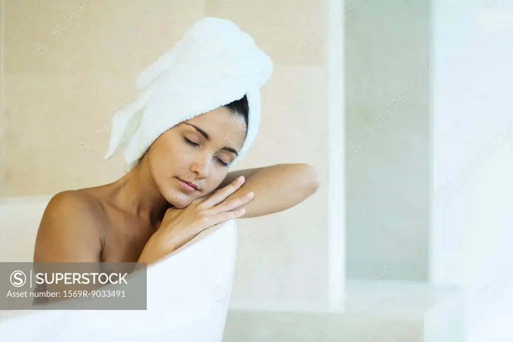 Woman sitting in bathtub with towel wrapped around hair, head resting on arms, eyes closed