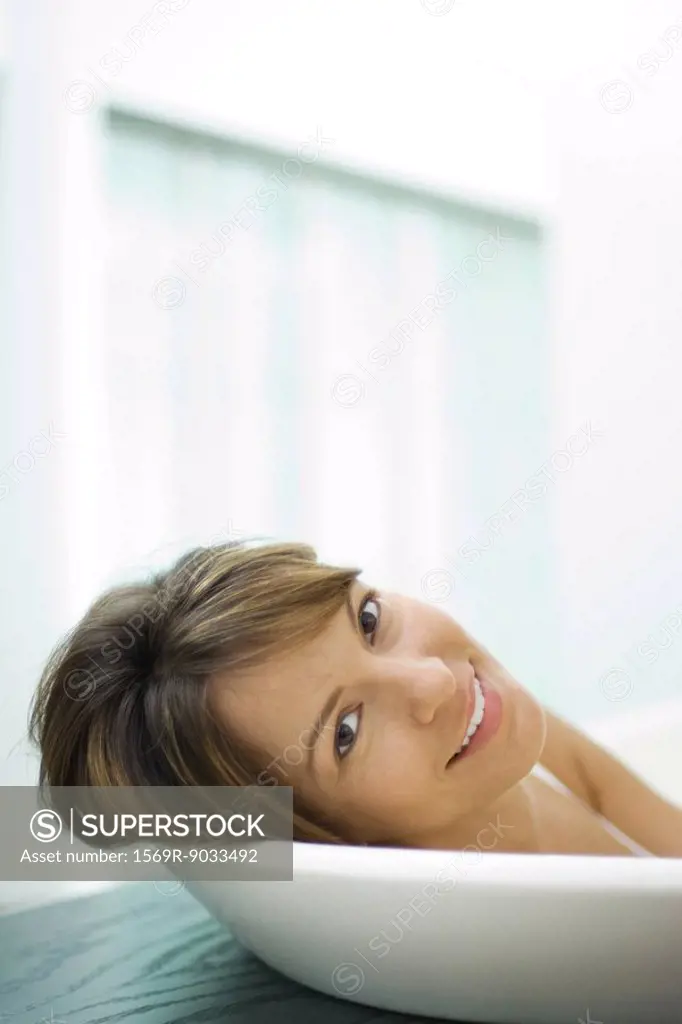 Woman leaning head against side of bathtub, smiling at camera, close-up