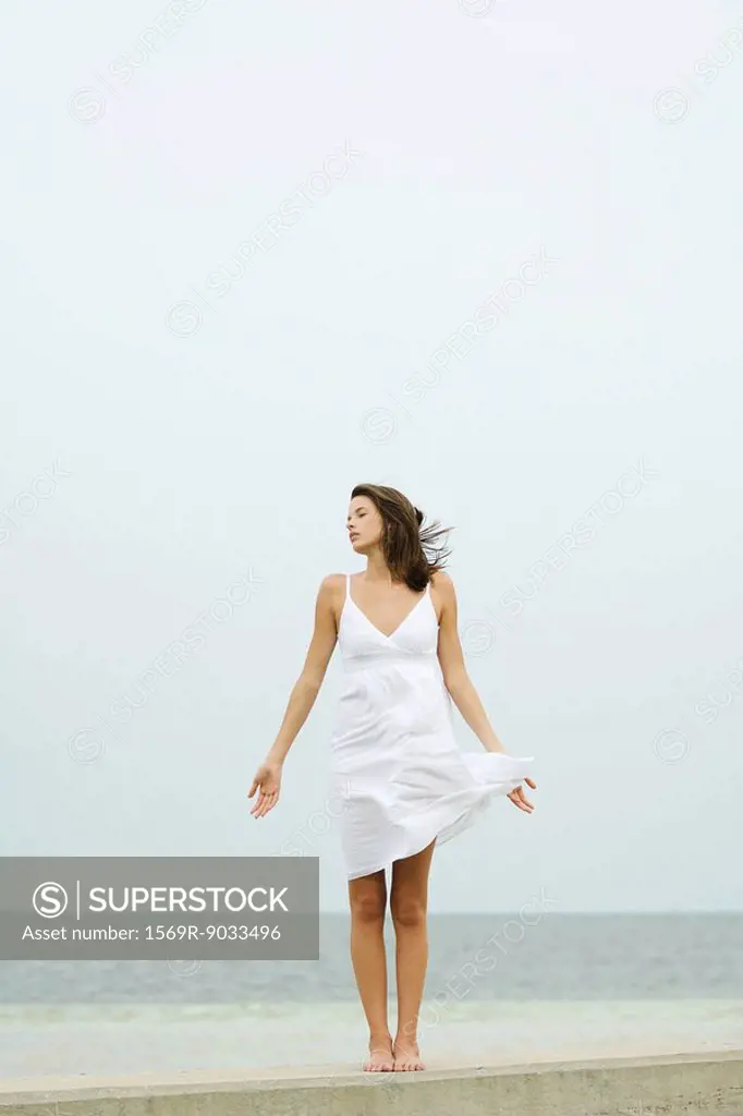 Teenage girl in sundress standing at the beach, tousled by wind, eyes closed