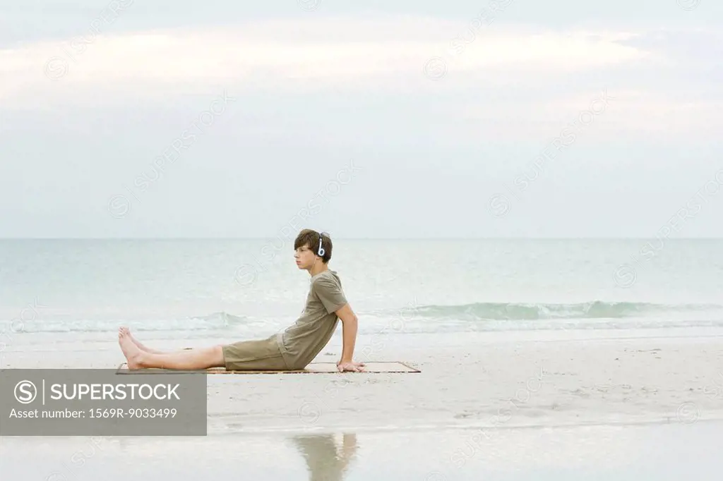 Teenage boy sitting on the beach, listening to headphones, side view