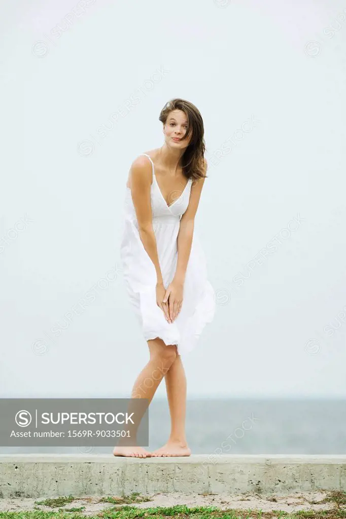 Teenage girl in front of the sea, bending over to hold skirt, tousled by wind, smiling at camera