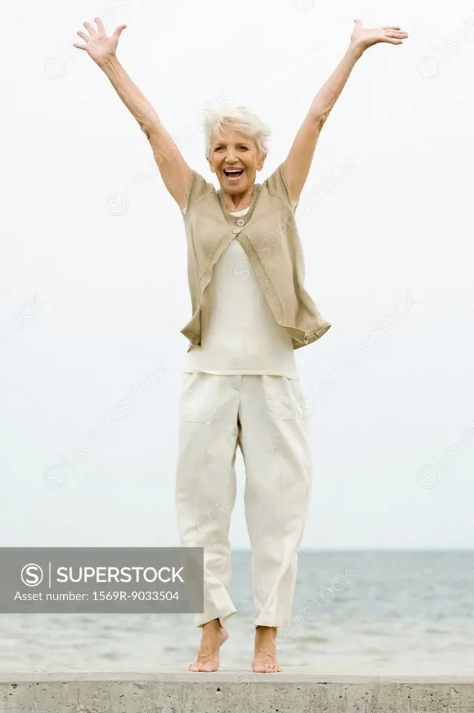 Senior woman standing on tiptoe at the beach, arms raised, smiling