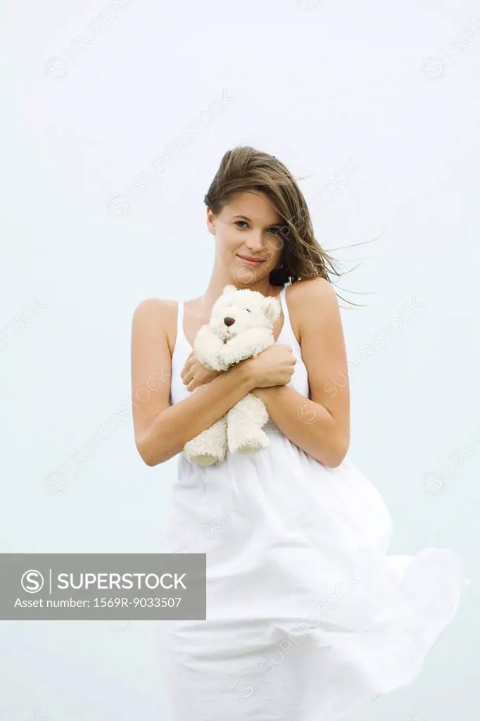 Teenage girl in sundress holding teddy bear, touseled by wind, smiling at camera