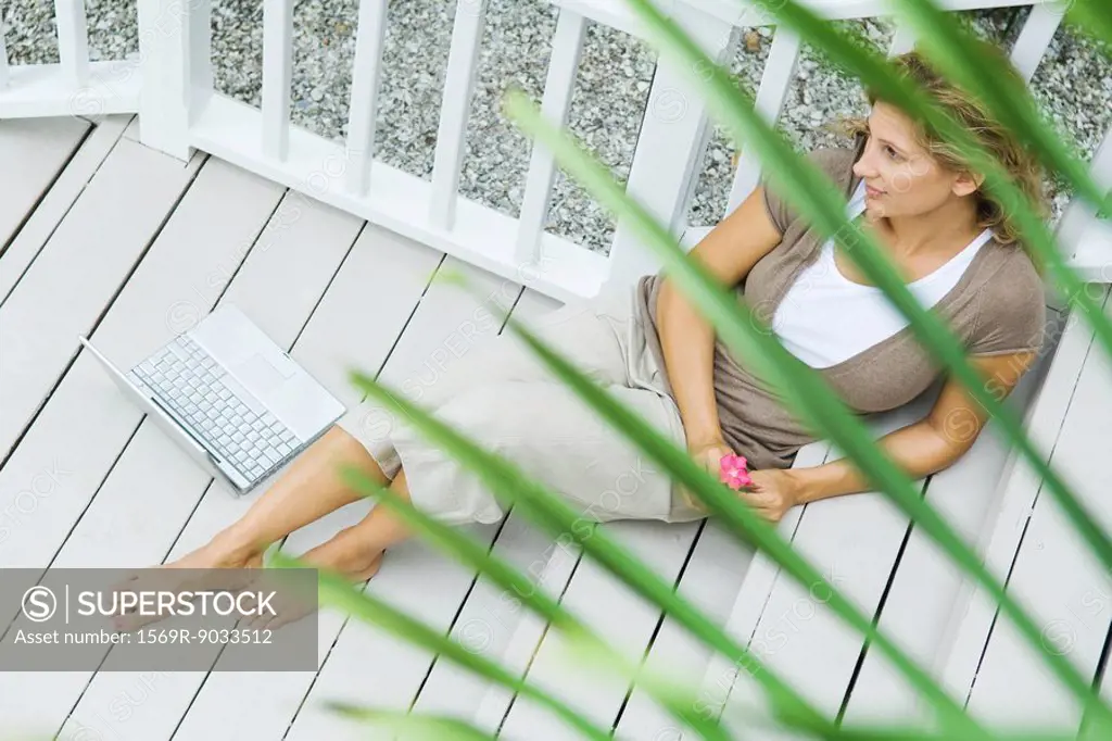 Woman sitting on porch next to laptop computer, holding flower, looking away, high angle view