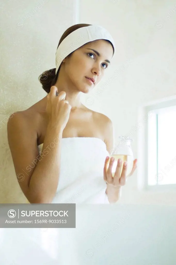 Young woman wrapped in towel applying perfume to neck, looking away