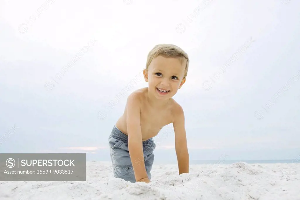 Little boy crouching at the beach, smiling at camera, portrait