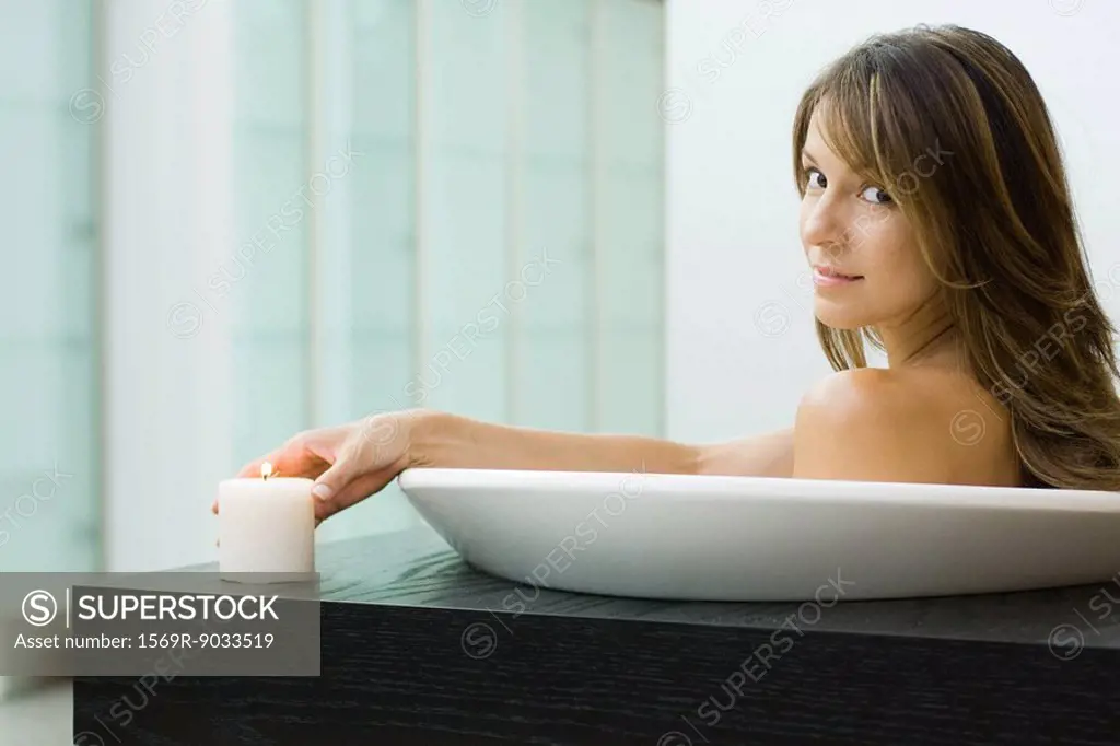 Woman sitting in bathtub, holding lit candle, smiling over shoulder at camera