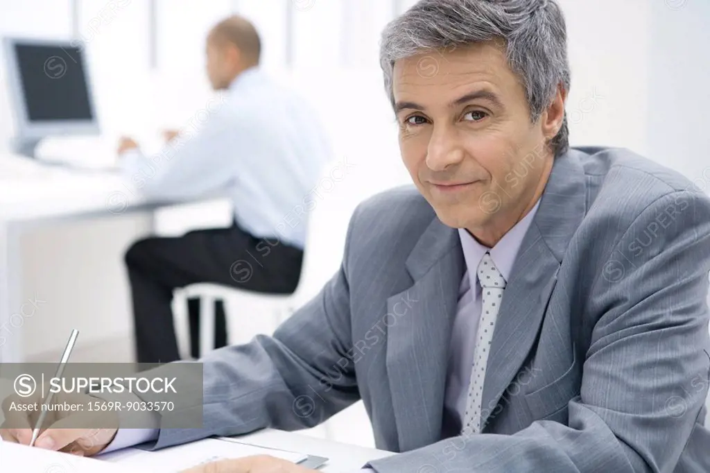 Businessman sitting at desk, using pen, smiling at camera