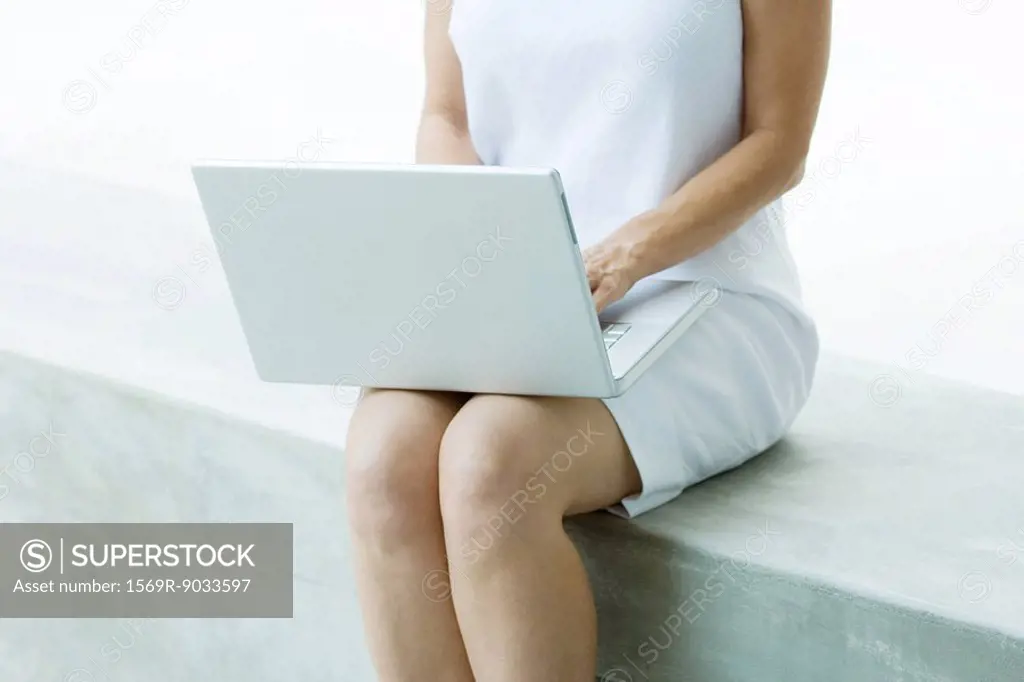 Woman sitting on low wall, using laptop computer, cropped view