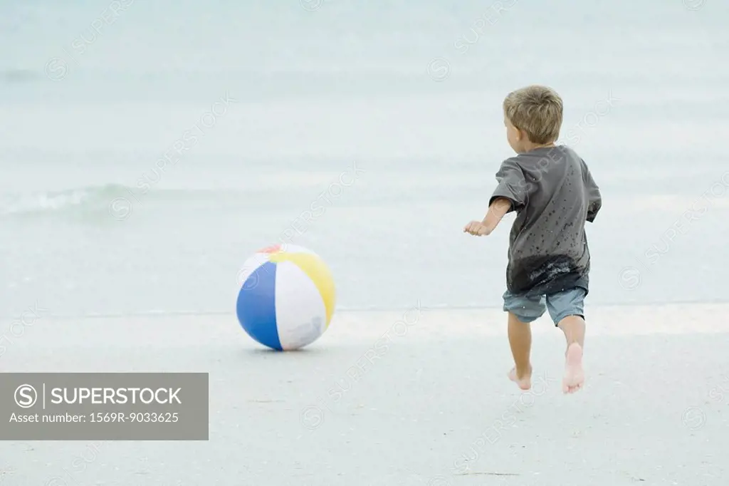 Young boy running after beach ball at the beach, rear view