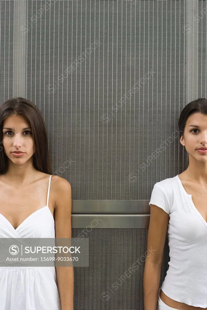 Teenage twin sisters standing in front of metal grate, both looking at camera, cropped