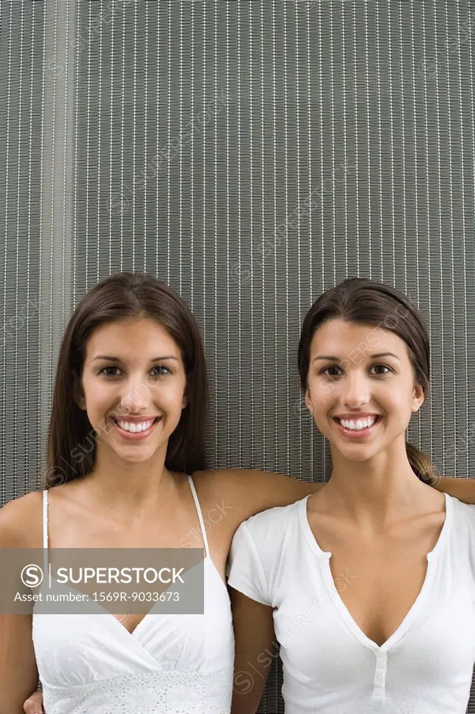 Teenage twin sisters with arms around one another, both smiling at camera, portrait