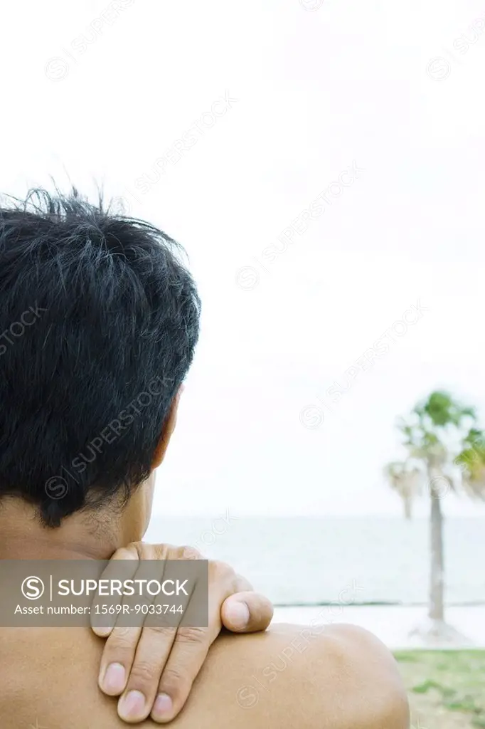 Man at the beach, hand on shoulder, cropped rear view