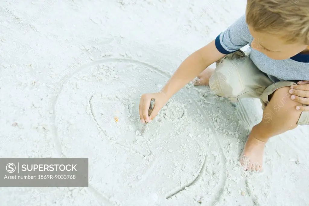 Little boy crouching at the beach, drawing in sand with stick, tilted view