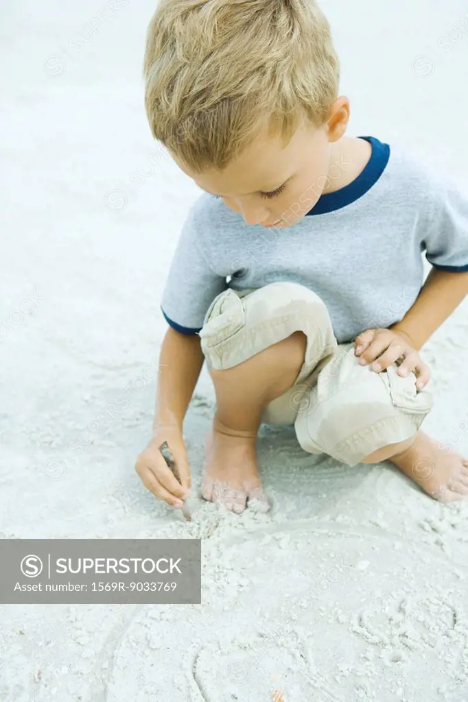 Little boy crouching at the beach, drawing in sand with stick