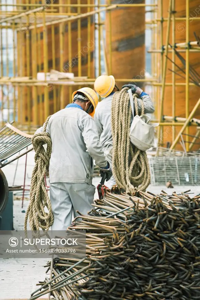 Workers at construction site carrying rope, metal rods stacked in foreground