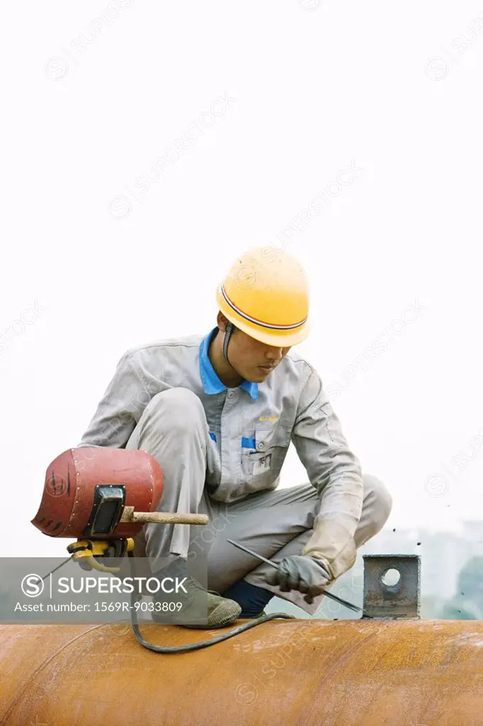 Worker crouching on metal pipe, holding welding mask, looking down