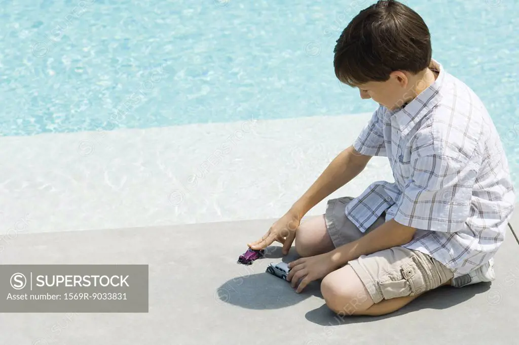 Boy sitting on ground, playing with toy cars, next to pool