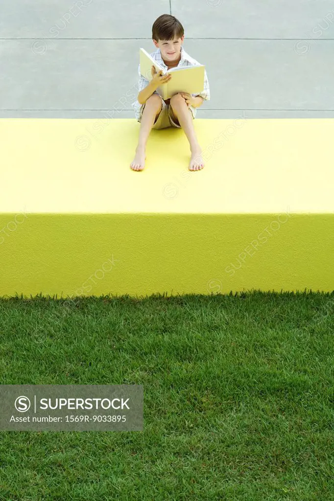 Boy sitting on low wall outdoors, holding book, looking away, high angle view