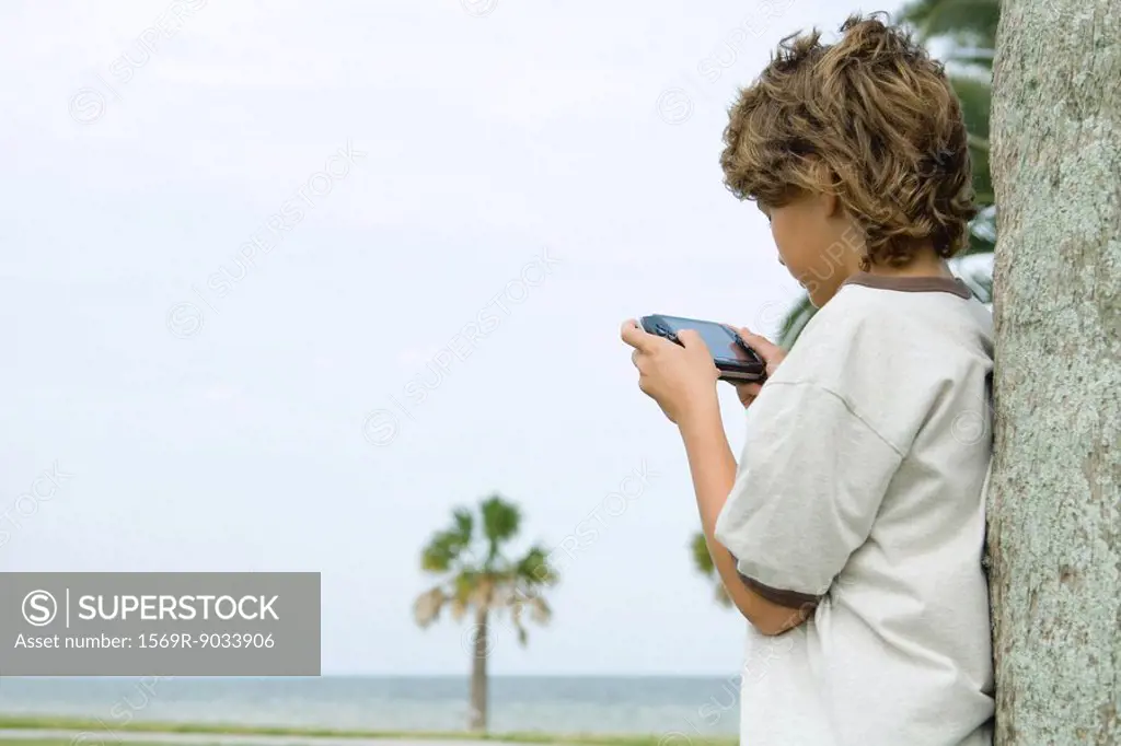 Boy leaning against tree trunk, playing handheld video game, side view