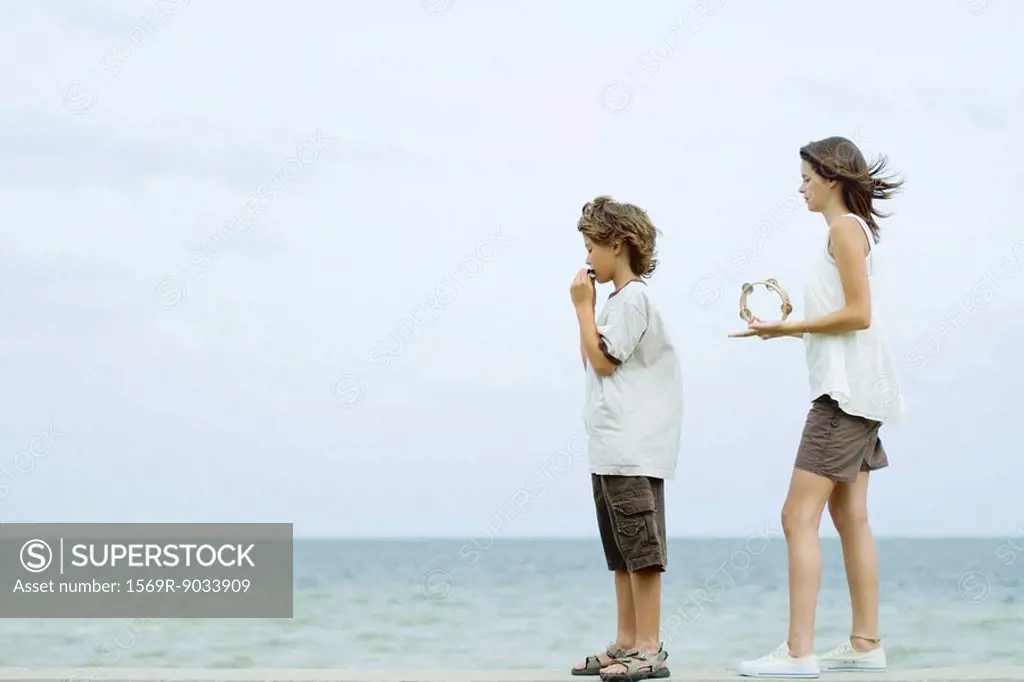 Siblings standing at the beach, boy playing harmonica, teen girl playing tambourine, looking at view