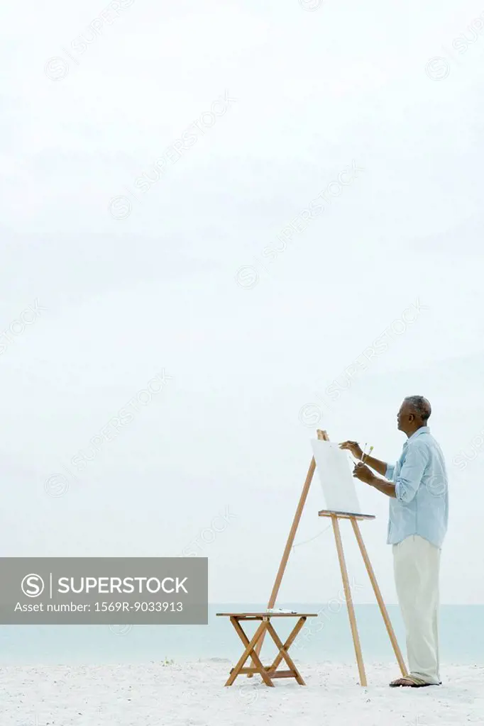 Senior man painting on canvas at the beach, side view