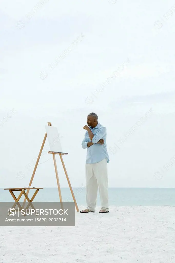 Senior man standing in front of blank canvas at the beach, holding paint brushes
