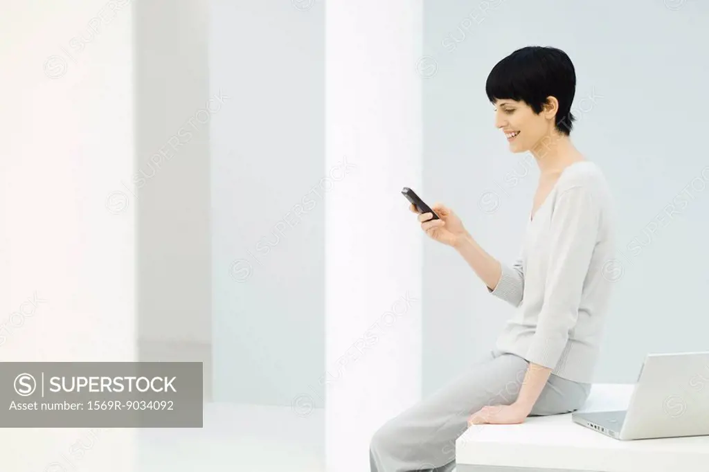 Woman sitting on edge of desk, looking at cell phone and smiling, side view