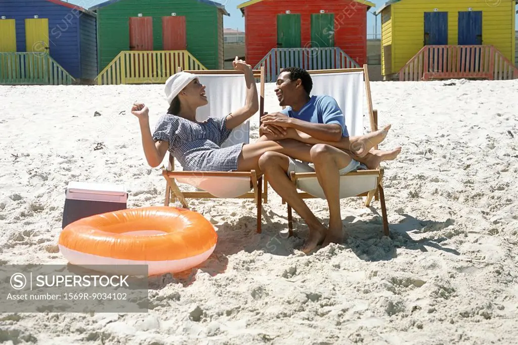 Couple sitting in lounge chairs at the beach, smiling, woman´s legs on man´s lap