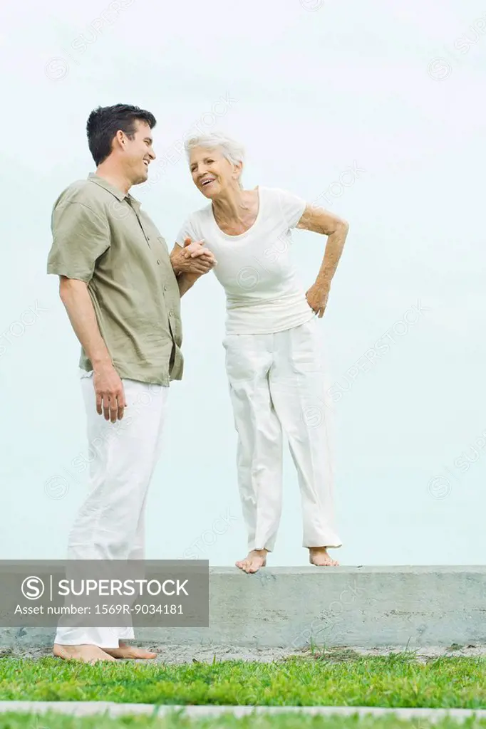 Senior woman standing on low wall next to adult son, holding hands, smiling