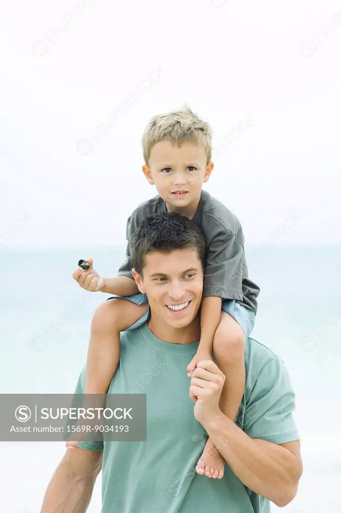 Man carrying son on his shoulders, boy holding seashell, both smiling