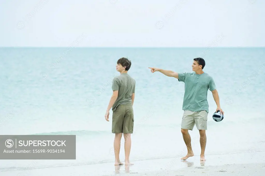 Father and teen son at the beach, man holding ball and pointing, both looking away