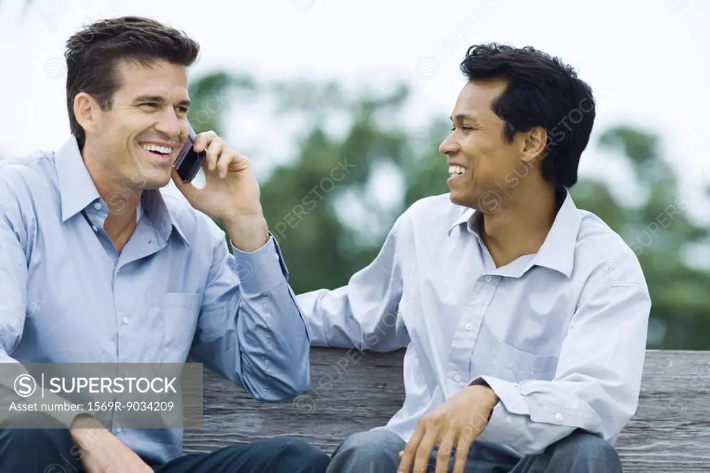 Two men sitting on bench, smiling, one using cell phone