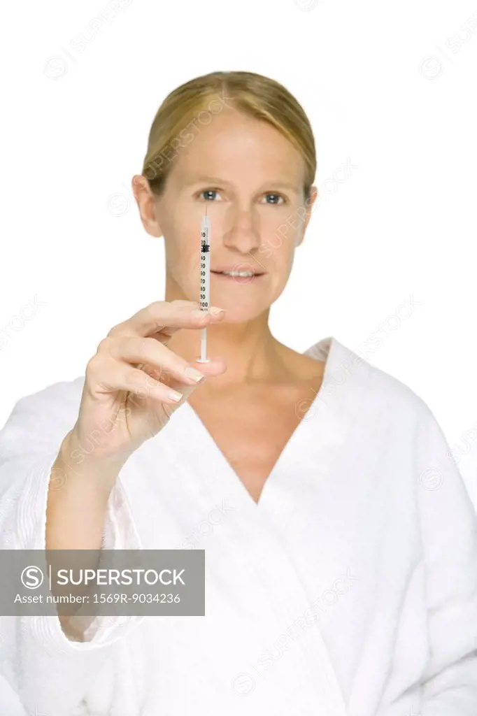 Woman in bathrobe holding up syringe, looking at camera