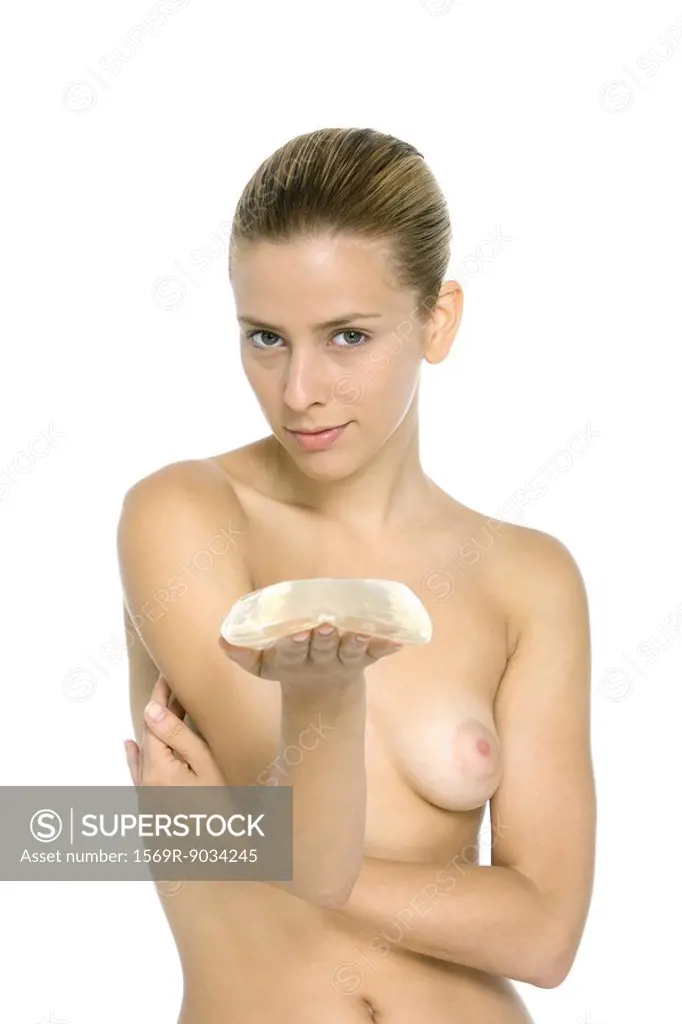 Nude woman holding breast implant, looking at camera