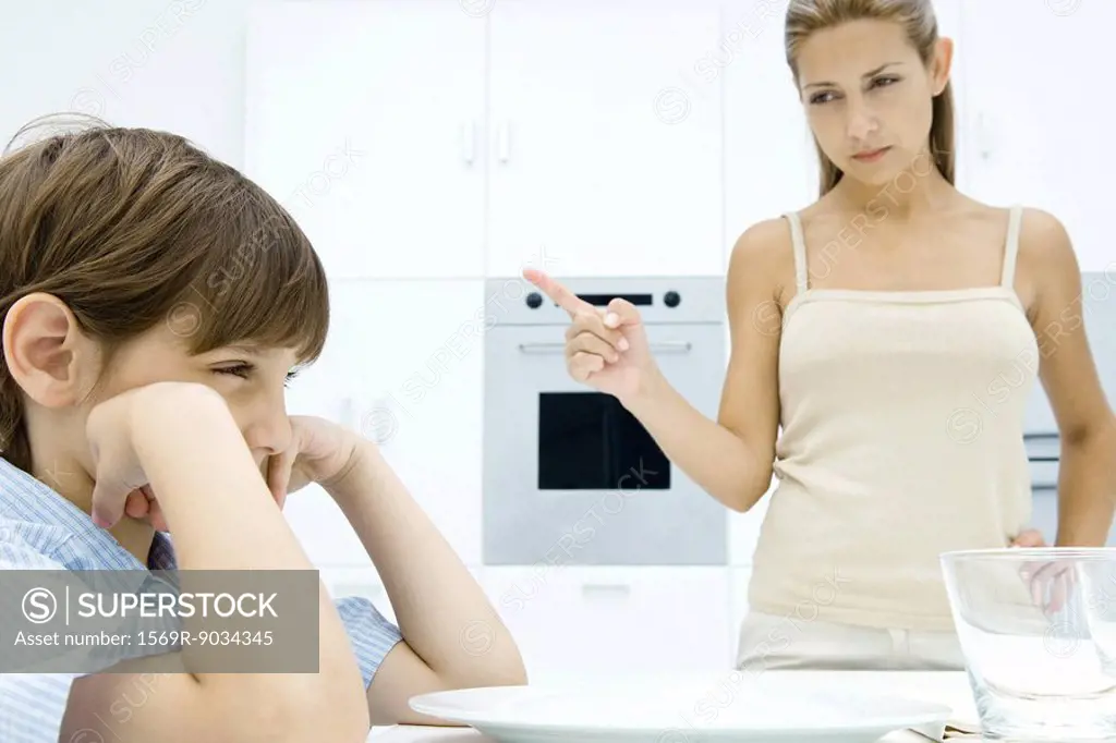 Boy pouting at kitchen table, mother shaking her finger