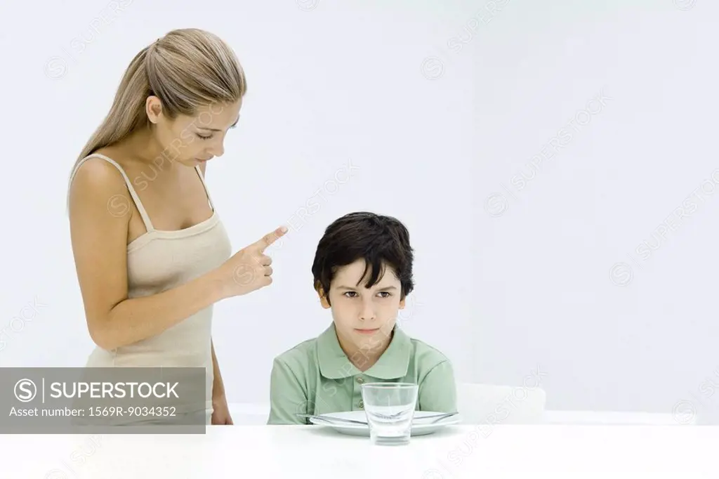 Boy sulking at table, mother standing beside him, shaking her finger