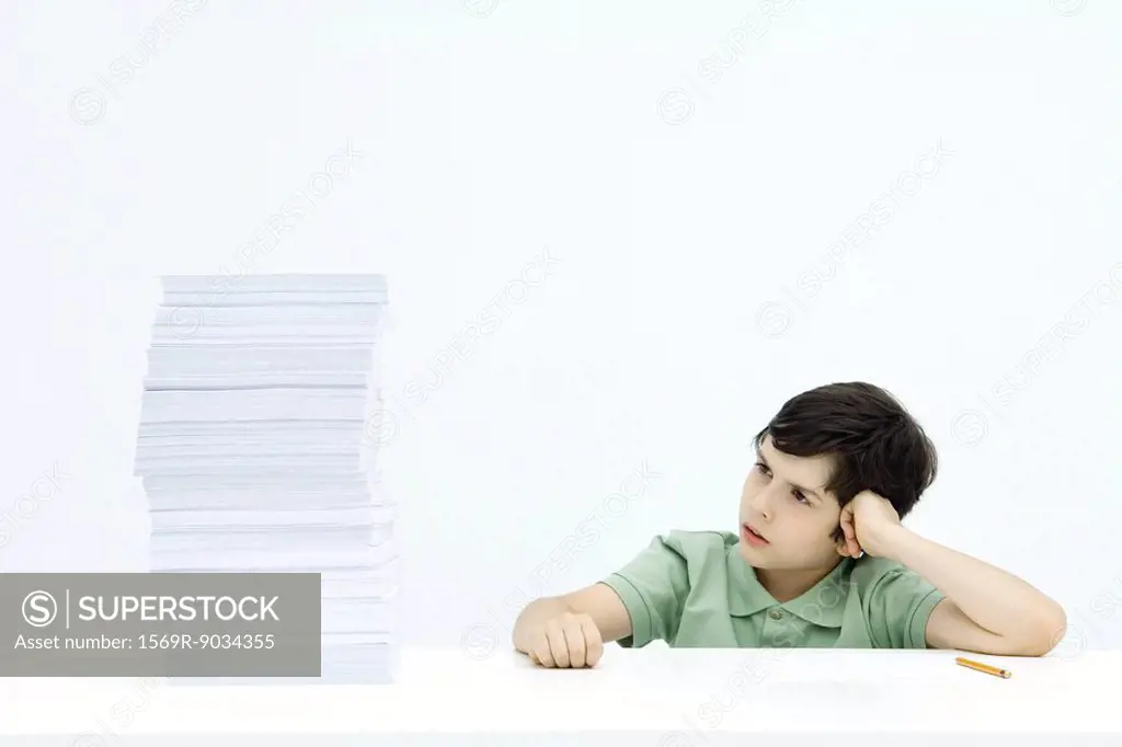 Boy sitting at table next to tall stack of homework, frowning