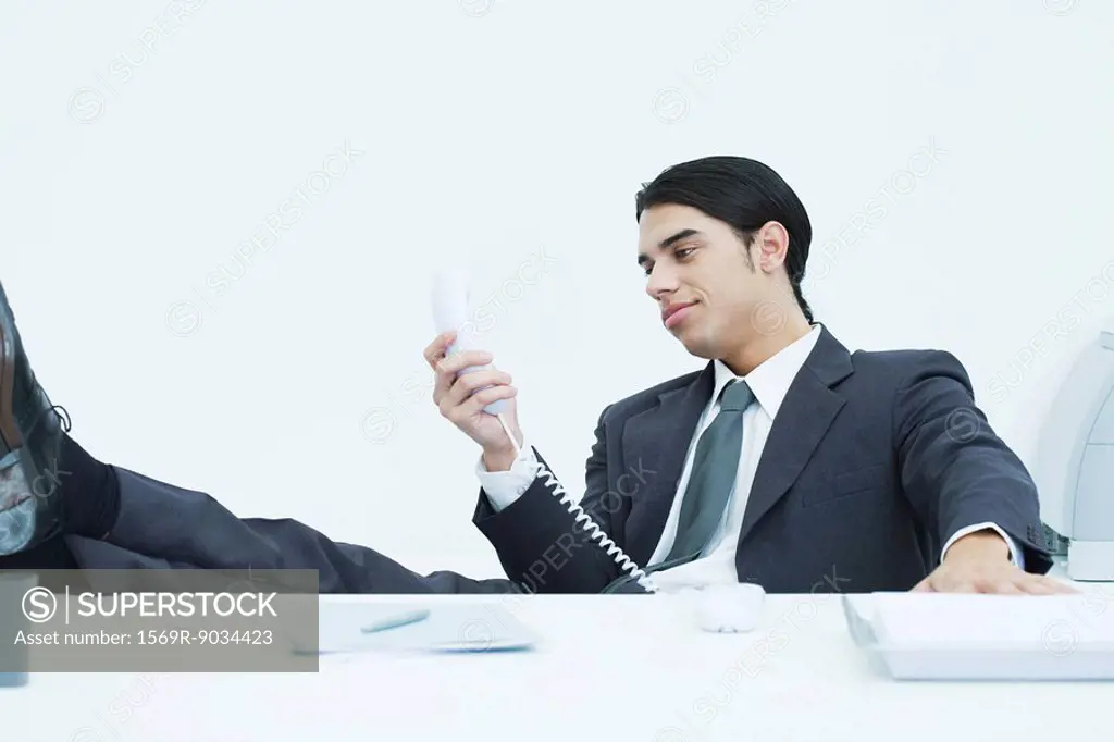 Young businessman sitting at desk with feet up, looking at landline phone, smiling