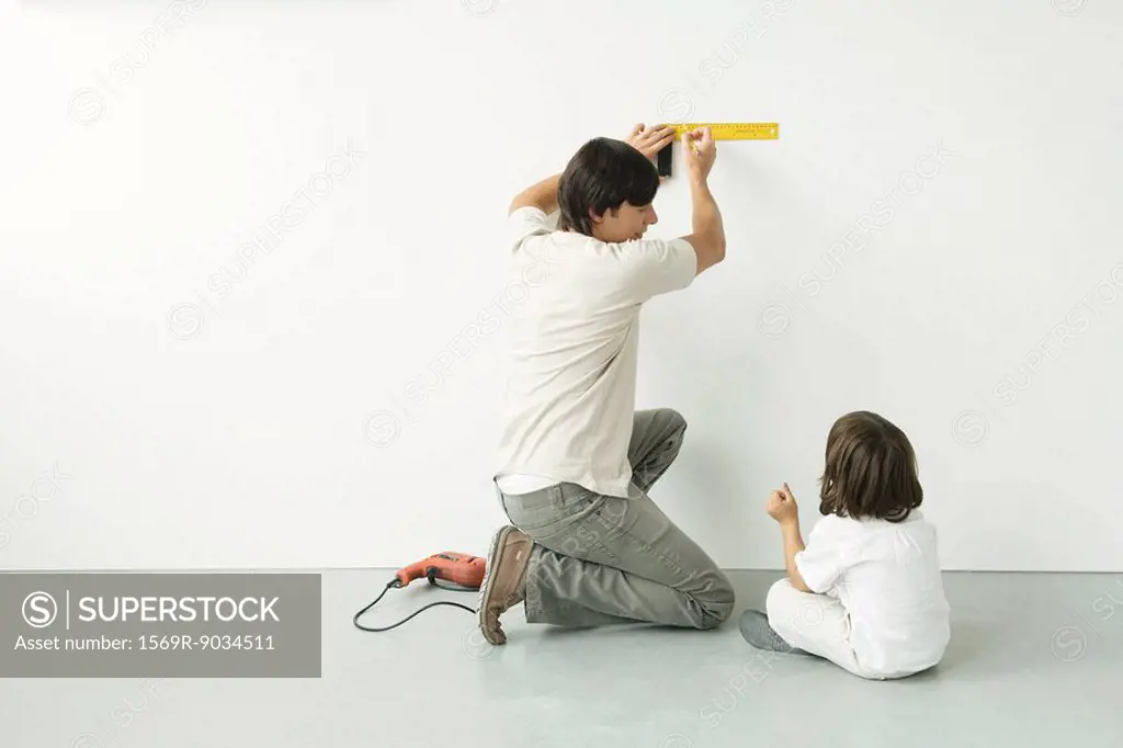 Man measuring wall with a ruler while his son watches, drill on the floor nearby