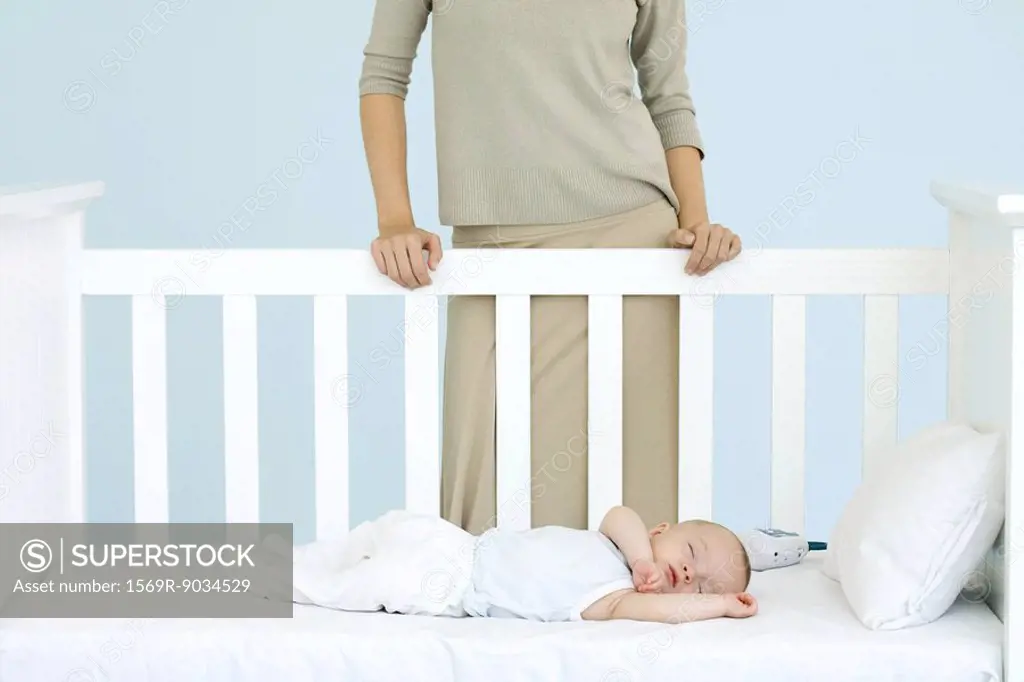 Mother standing behind crib as infant sleeps, cropped view