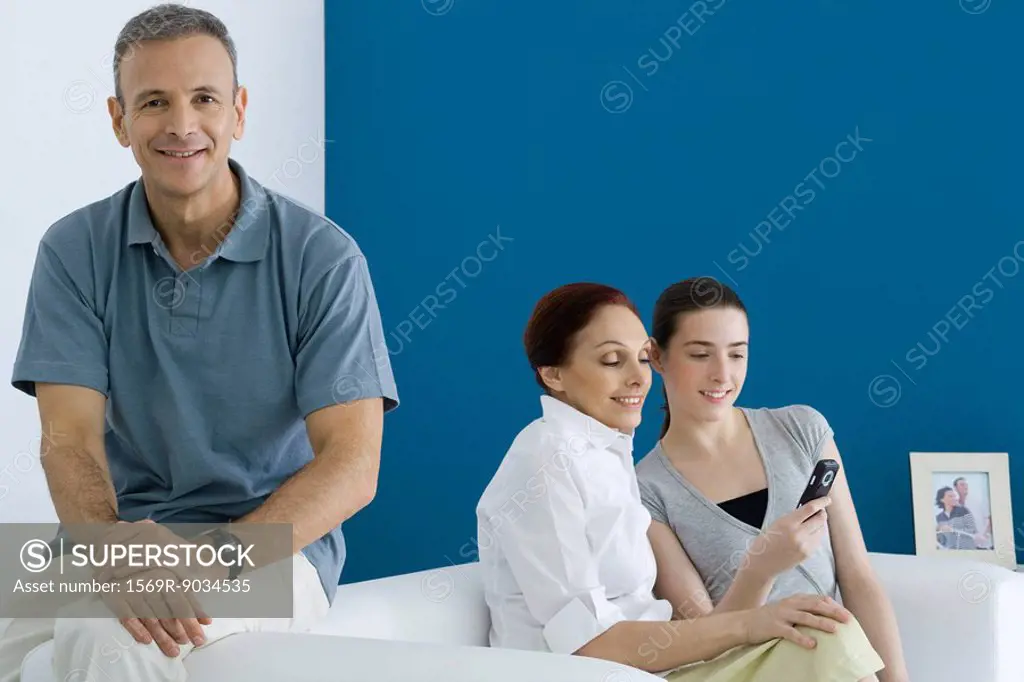 Man sitting on sofa, smiling at camera, his wife and daughter looking at cell phone behind him