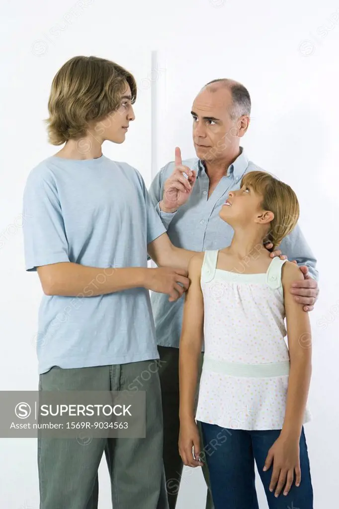 Father shaking his finger at teen son, preteen daughter watching and smiling