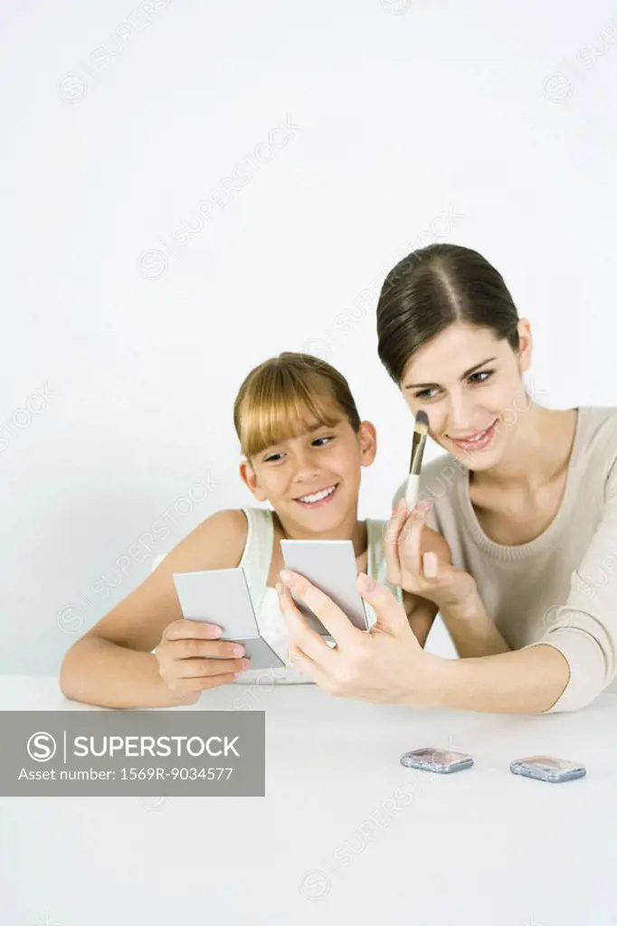 Young woman sitting next to preteen sister, putting on make-up, both looking into hand mirrors
