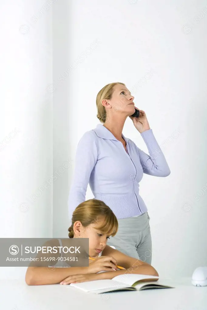 Preteen girl sitting at table with homework, mother standing beside her, using cell phone
